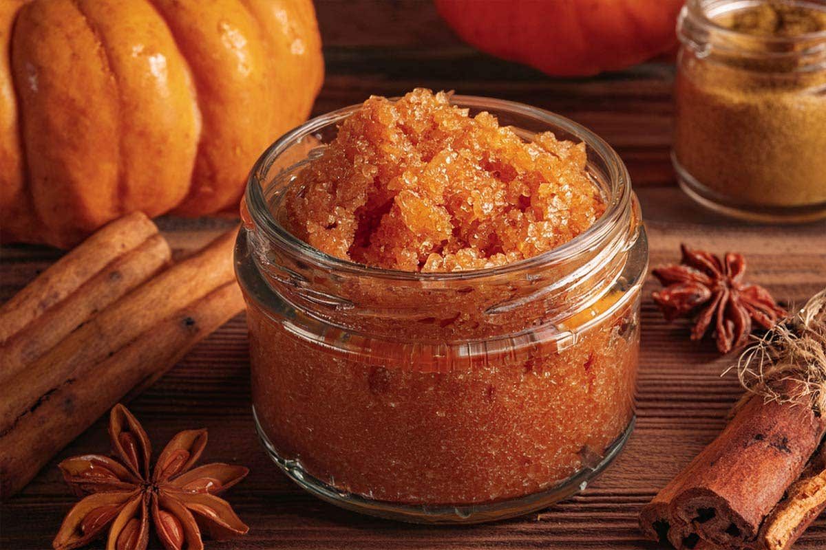 A small glass jar full of an orange-colored sugar scrub sits on a wood table surrounded by spices and pumpkins.