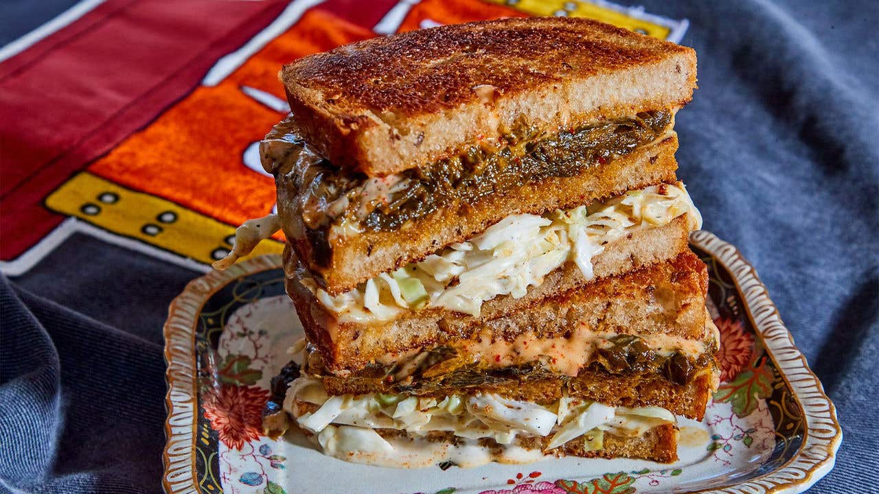 Two halves of a sandwiched stacked on top of each other, sandwich has collard greens, col slaw and sauce. 