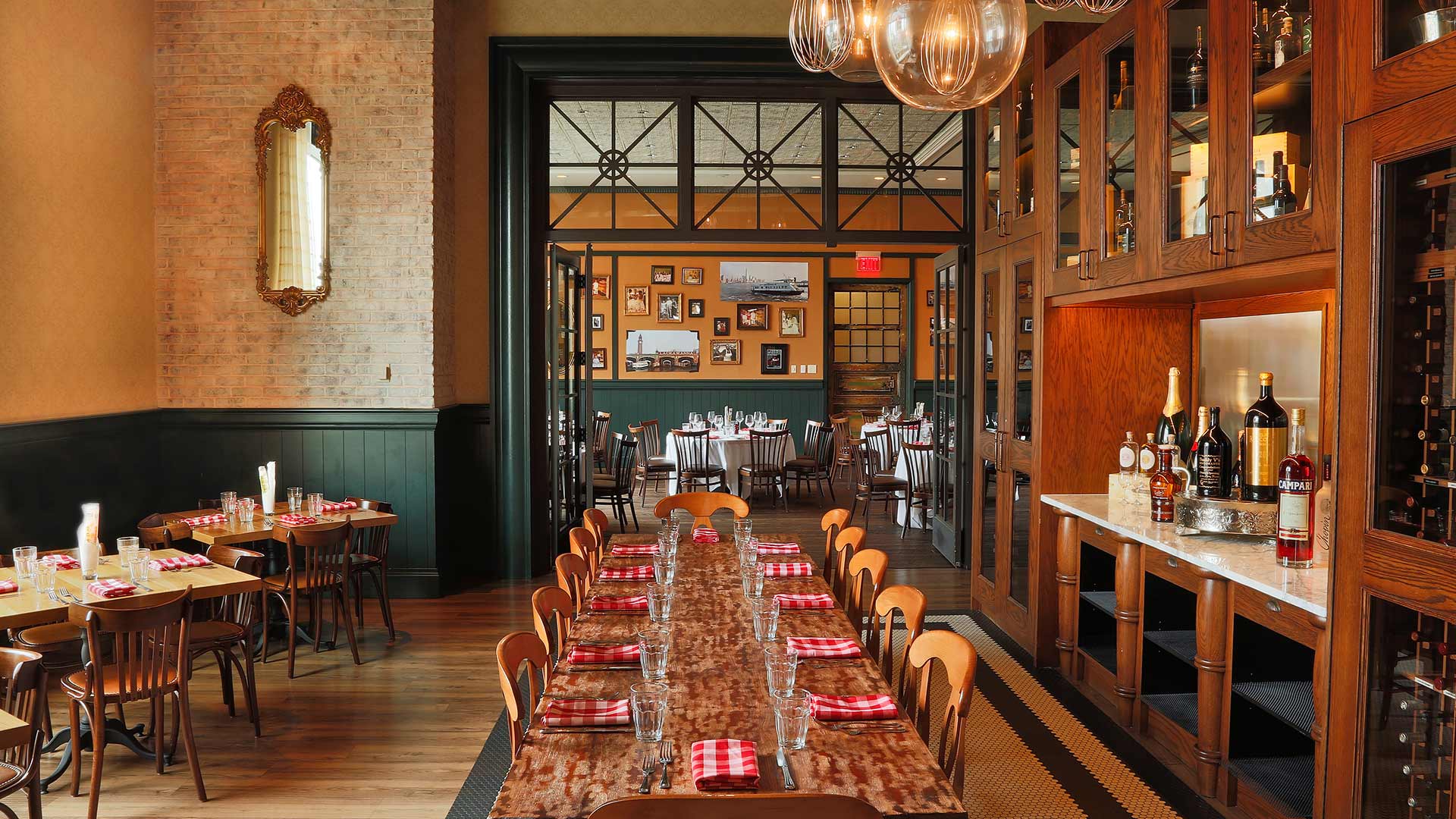 A cozy restaurant dining area with wooden tables, chairs, and pendant lighting, featuring a wine display and framed photos on walls.