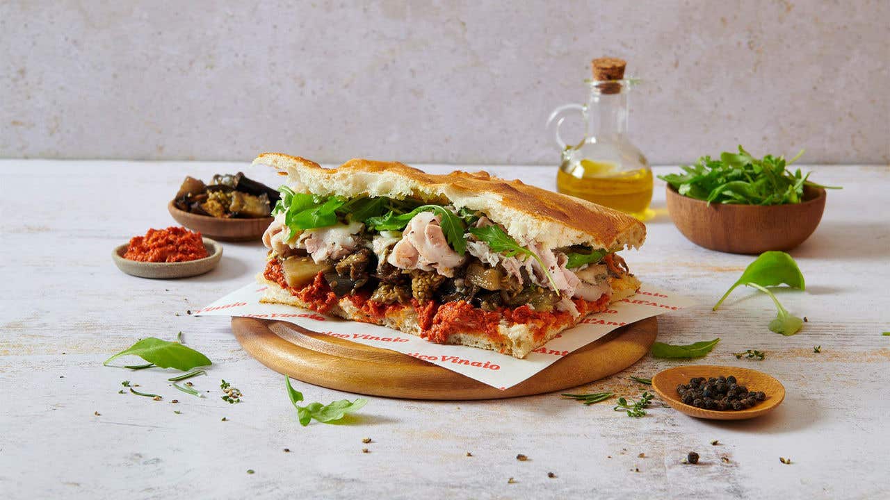 Panini sandwich with turkey, lettuce, olives and sun dried tomatoes on a round wood plate surrounded by seasonings and a bottle of olive oil. 