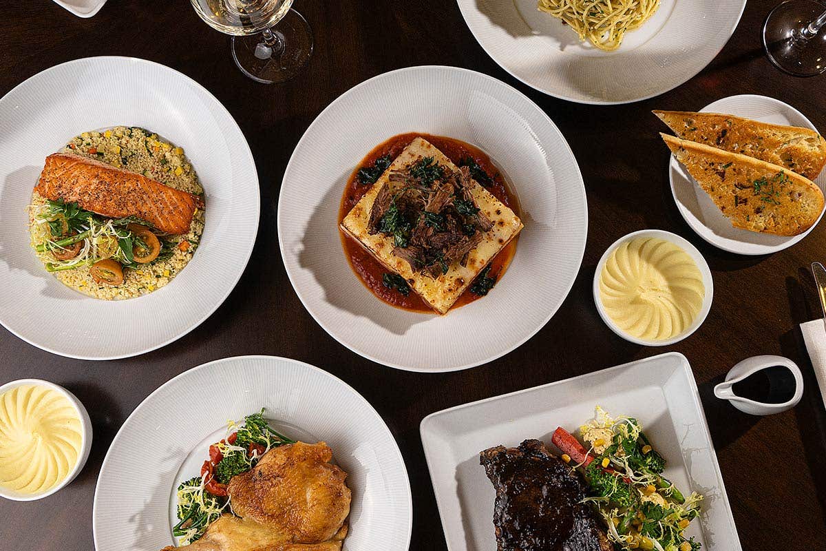 A table set with plated entrées including grilled salmon on rice, braised short rib lasagna, roast chicken, steak, pasta, garlic bread, mashed potatoes, and white wine.