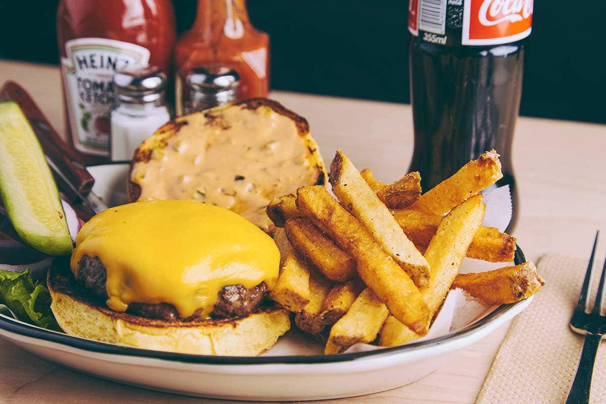 Cheeseburger with fries on a plate, accompanied by ketchup, hot sauce, and a bottle of Coca-Cola.