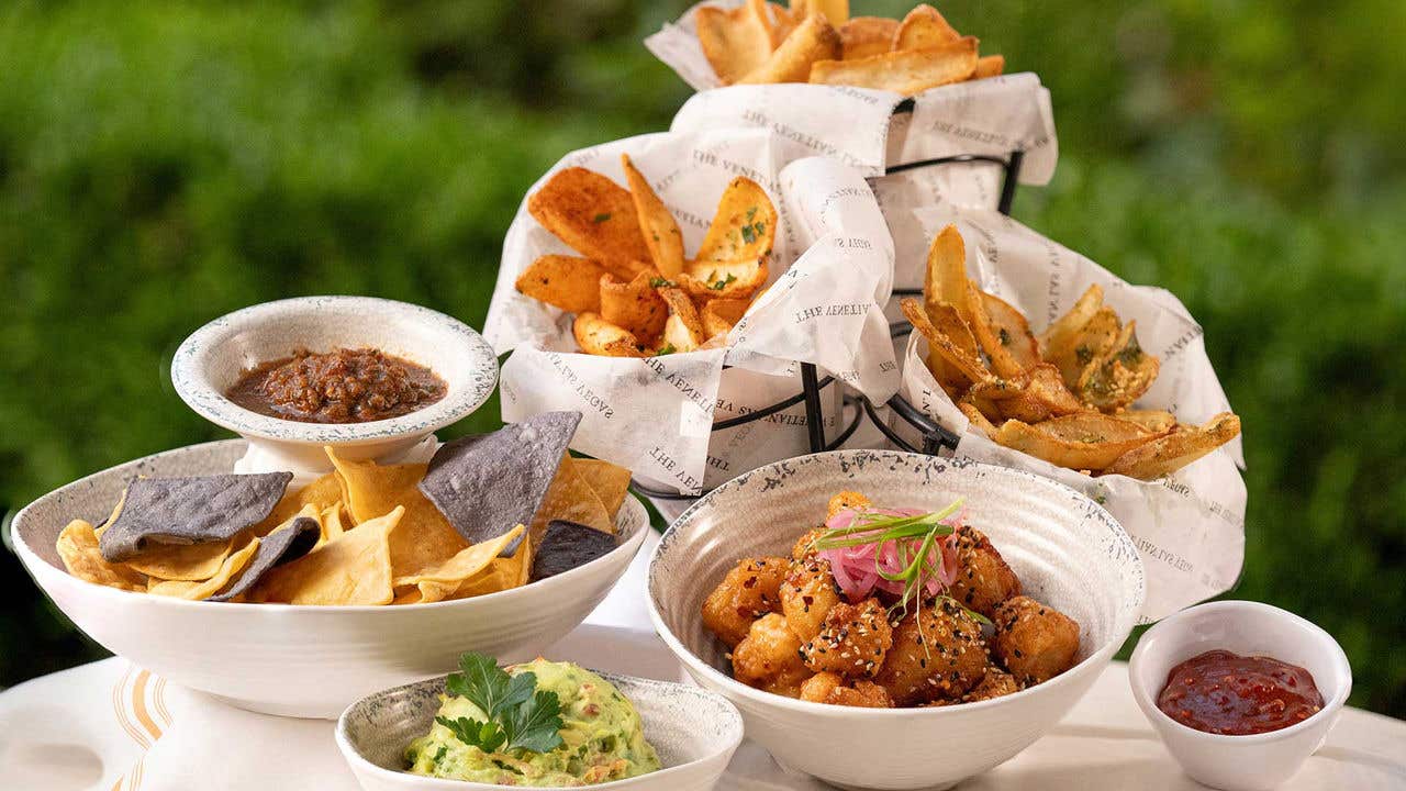 Seven bowls of appetizers sit on a marble table with green bushes in the background.