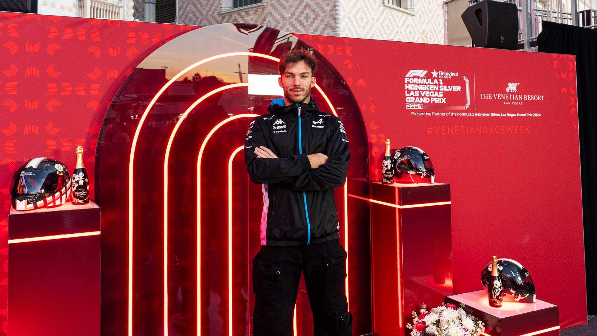 A race car driver crossing his arms in front of a photo platform that primarily has a U shaped lights behind red stained glass material, alongside race helmets and bottles of champagne