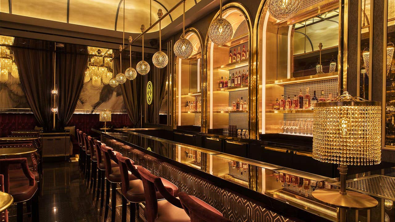 A long angle of a gold toned bar with burgundy bar stools and hanging globe lights.